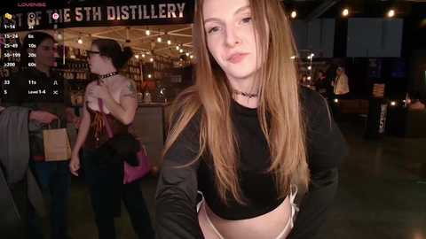 Media: Video of a young, pale-skinned, long-haired woman in a black crop top, standing in a dimly lit bar with wooden shelves, wearing a black choker.