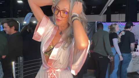 Video of a middle-aged woman with gray hair, wearing glasses and a pink satin robe, posing at a crowded event.