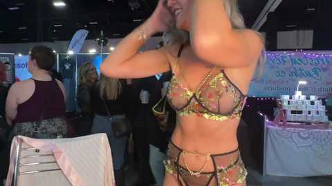 Video of a blonde woman in a sheer, floral-patterned bra and panties, adjusting her hair at an event; background shows other attendees, banners, and merchandise tables.