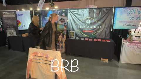 Video of a trade show booth featuring two women in black jackets, one with long blonde hair, displaying products; background includes banners and product displays.
