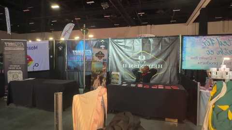 Video of an indoor exhibition booth with two people at a table. Displays include a TV screen, banners, and promotional materials.