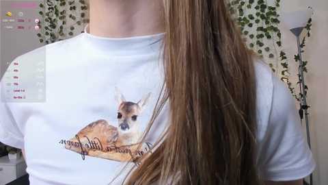 Video of a person with long, straight brown hair wearing a white T-shirt with a graphic of a fawn lying on a log. The background shows a room with a green curtain and a floor lamp.