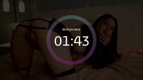 Video of a nude woman in black bondage gear, bent over a bed, with a dimly lit, shadowy bedroom background.