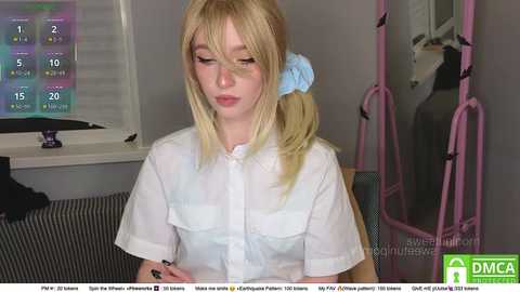 A video of a young, fair-skinned woman with blonde hair in pigtails, wearing a white blouse, sitting indoors with a pink exercise machine and window with blinds in the background.