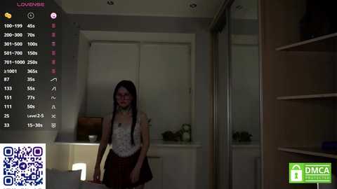 Video of a young woman with long black hair, wearing a white lace top and brown skirt, standing in a dimly lit bedroom with a large mirror and shelves.