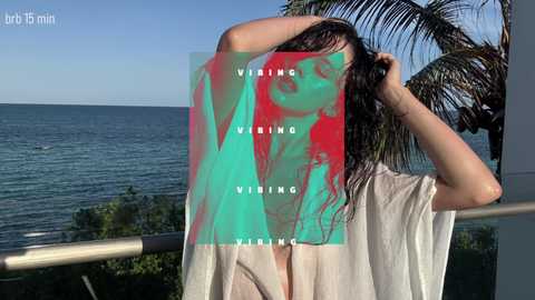 A video of a woman with long, wet hair, wearing a white robe, standing on a balcony with a palm tree and ocean in the background. Text overlay reads \"VIRING VIENING\" and \"art 15 min.\