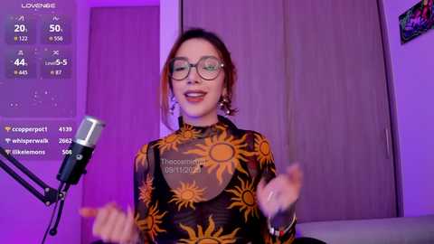 Video of a smiling, light-skinned woman with glasses and orange floral top, standing in front of a purple-lit room, holding a microphone.