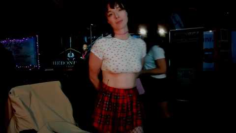 A video of a young woman with short brown hair, wearing a white polka-dot crop top and a red plaid skirt, standing in a dimly lit bar with neon signs and a television screen in the background.