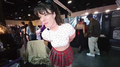 Video of a woman with short dark hair, wearing a white polka-dot crop top and red plaid skirt, standing in a dimly lit, crowded indoor market.