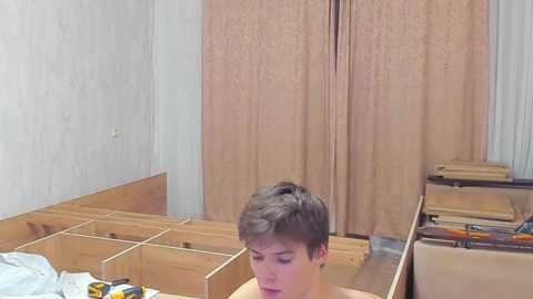 Video of a young, bare-chested man with short brown hair, sitting in a room with beige carpet, partially constructed wooden shelves, and beige curtains.