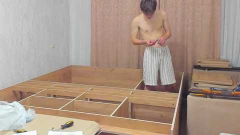 Video of a shirtless, slender man with short brown hair wearing striped pajama shorts, standing in a workshop with a wooden frame, tools, and beige curtains.