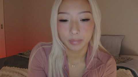 Video of a young Asian woman with long, straight platinum blonde hair, light skin, wearing a pink long-sleeve top, in a dimly lit bedroom with beige walls and a bed with patterned sheets.