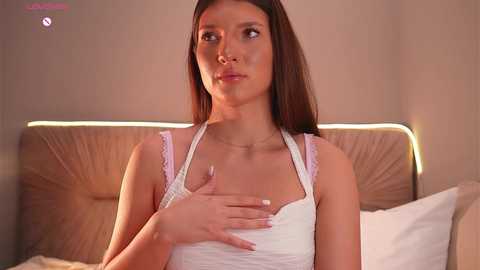 Video of a young, fair-skinned woman with long brown hair, wearing a white lace camisole, touching her chest in a dimly-lit bedroom with beige walls and a cushioned headboard.
