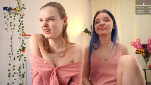 Video of two young Caucasian women with fair skin, one with brown hair, the other with blue hair, wearing off-shoulder pink tops, smiling. Background features floral decor, pink flowers, and a lamp.