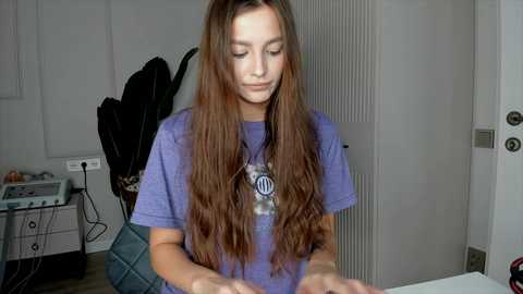 Video of a young woman with long, wavy brown hair, wearing a purple T-shirt, sitting at a table, eyes closed, focused. Background shows a black guitar, white wall, and a door.
