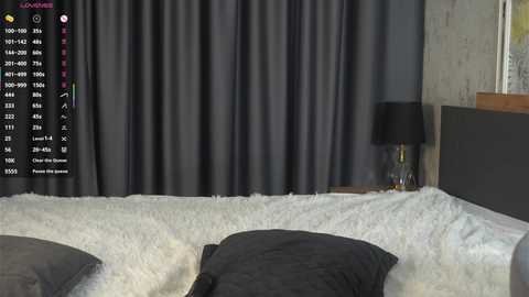 Video of a modern bedroom with a grey bed, dark curtains, white fluffy rug, black lamp on a nightstand, and a digital screen overlay displaying \"Lovense\" controls.