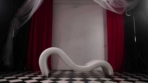 Video of a sleek, white, curvaceous chaise lounge with a flowing design, set against a backdrop of black-and-white checkered floor, red curtains, and sheer white drapes in a dimly lit room.
