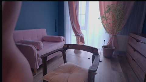 Video of a pastel-colored, minimalist living room with a pink sofa, chair, and sheer curtains; a tall plant stands in the corner.