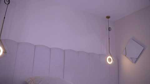 Video of a minimalist, modern bedroom with a light purple wall, white bedding, and three pendant lights with circular bulbs. The room exudes a serene, monochromatic vibe.