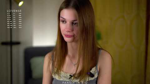 Media: Video of a young woman with straight, light brown hair, wearing a gray tank top with a floral pattern. She has a fair complexion and is indoors, with a blurred yellow and white wallpaper background.