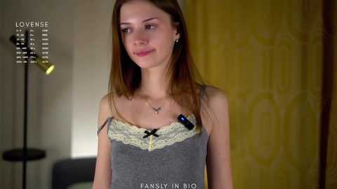Video of a young Caucasian woman with straight brown hair, wearing a gray tank top with yellow lace trim, standing indoors with a yellow-patterned curtain in the background.