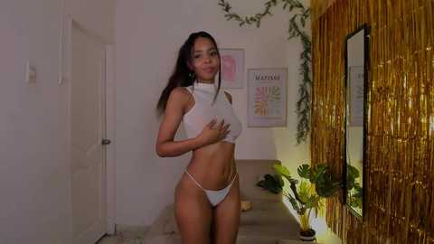 Video of a slender, tan-skinned woman with long dark hair in white lingerie, standing in a dimly lit, modern, bamboo-walled room with potted plants and framed art on the wall.
