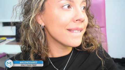 Video of a smiling woman with curly, shoulder-length hair, wearing a black top, and a thin silver necklace. Background shows a purple chair and a cluttered desk. Text at the bottom reads \"@alonessarez OnlyFans OnlyFans free.\