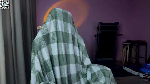 A video showing a green and white checkered blanket draped over a chair, with a soft, warm glow from an unseen source creating a halo effect. The background features a pink wall, exercise equipment, and a window with gray curtains.