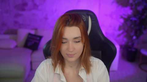 Media: Video of a young woman with straight, shoulder-length auburn hair, wearing a white blouse, seated in a black gaming chair. Background features a dimly lit room with purple lighting and a leafy plant.
