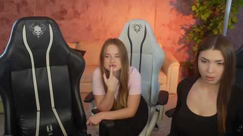 Video of three young women in a gaming setup. Two sit in black gaming chairs with white accents, one with long brown hair, one with blonde hair. The third woman stands in the background with brown hair, all wearing casual clothes.
