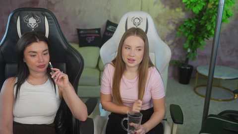 Video of two young women in gaming chairs, one with a white tank top, the other in a pink t-shirt, holding controllers, in a modern, minimalistic room with a green plant.