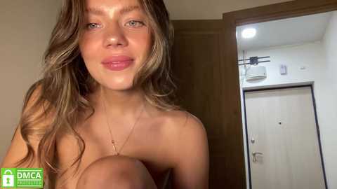 Media: Video of a smiling young woman with long, wavy brown hair, fair skin, and small breasts, standing nude in a bathroom.