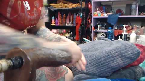 Video of a tattooed arm using a vibrating sex toy on a person's back in a cluttered room filled with various sex toys and paraphernalia.
