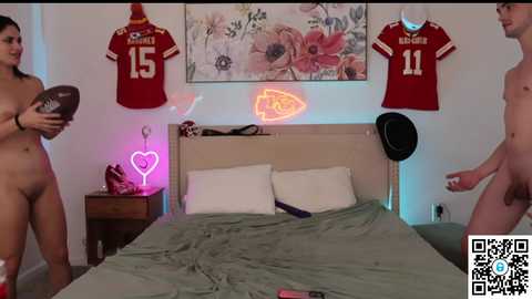 Video of a bedroom with a bed, two naked people, one holding a football, floral wall art, red jerseys, neon signs, and a QR code.