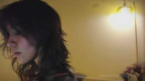 A video of a young person with shoulder-length dark hair, partially illuminated by a yellow light, standing in a dimly lit room with a beige wall and a small lamp.