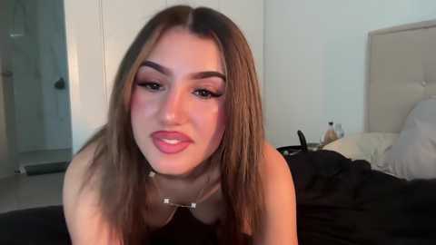 Video of a young woman with long brown hair, light skin, and full makeup, wearing a black top, in a modern bedroom with a beige headboard and messy bed.