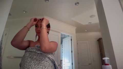 A video of a woman in a bathroom, wrapping a towel around her head, wearing a gray patterned dress.