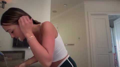 Video of a young Caucasian woman with light skin, brown hair, and a slim build, wearing a white tank top and green shorts, sitting in a modern, minimalist kitchen, looking at her phone.