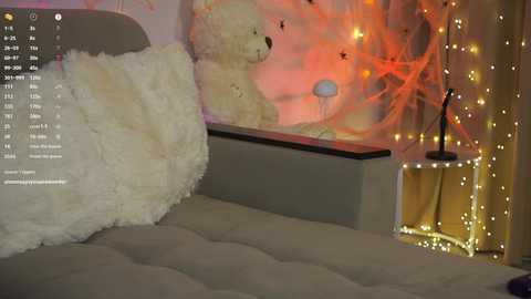 A video of a cozy bedroom with a beige bed, a plush white throw pillow, a teddy bear, and warm, glowing fairy lights on a wall.