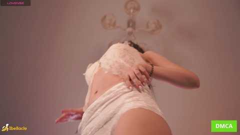 A video shows a woman with light skin, wearing white lace lingerie, adjusting her bra. The background features a beige ceiling with a chandelier. The image has a watermark from \"NubileFilms.\