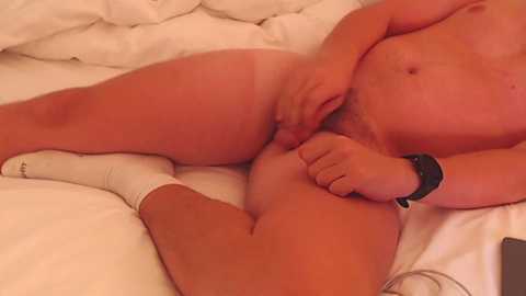 A video of a nude, fair-skinned man lying on a bed with white sheets, wearing a black smartwatch on his left wrist. His right hand is between his legs, revealing his erect penis.