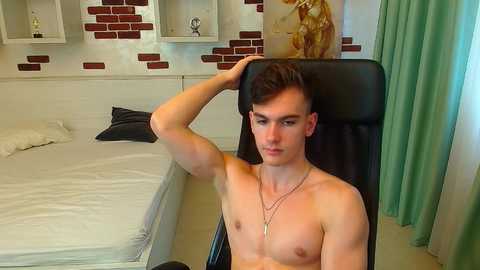Media: Video of a shirtless, young, muscular, fair-skinned man with short brown hair, sitting in a black gaming chair, wearing a necklace, in a modern, minimalist bedroom with white walls, brick-patterned accents, and teal curtains.