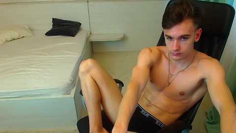Video of a shirtless, fit young man with short dark hair, wearing black Calvin Klein boxers, sitting on a black chair in a minimalist, white-walled room with a bed and pillow.
