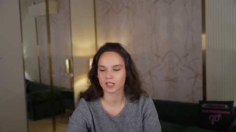 A video of a woman with medium-length wavy black hair, wearing a gray sweater, standing in a dimly lit room with marble walls and a green couch.