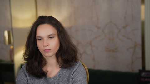 Video of a young woman with long, dark hair, wearing a grey sweater, sitting indoors against a marble wall with gold accents.