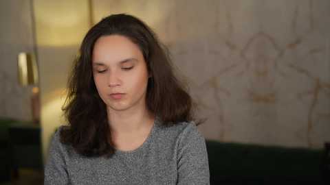 Video of a young woman with long, dark hair, wearing a gray knit sweater, sitting on a green couch in a softly lit, beige wallpapered room.