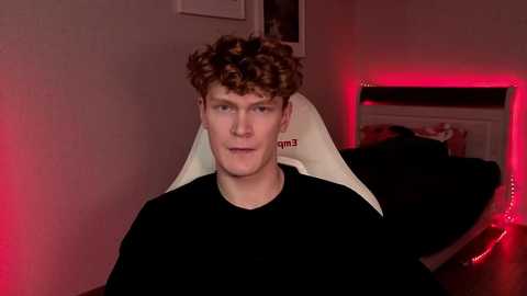 Video of a young man with curly brown hair, wearing a black shirt, sitting on a white gaming chair, in a dimly lit room with red LED strip lights, and a bed in the background.
