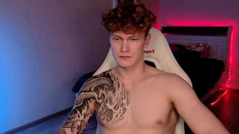 Video of a shirtless, pale-skinned young man with curly brown hair, sitting in a gaming chair. His left arm is heavily tattooed with a detailed snake design. Background features a dimly lit room with red and blue lighting, a bed, and a TV.