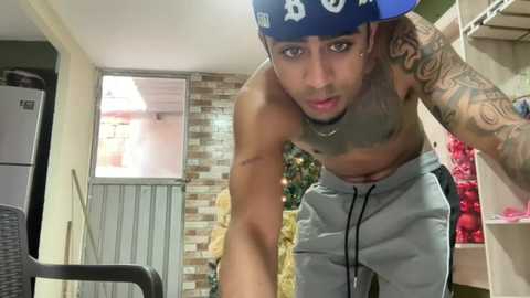 Video of a shirtless, tattooed Black man with a blue baseball cap, leaning over a chair in a modern kitchen with white cabinets and a brick wall background.