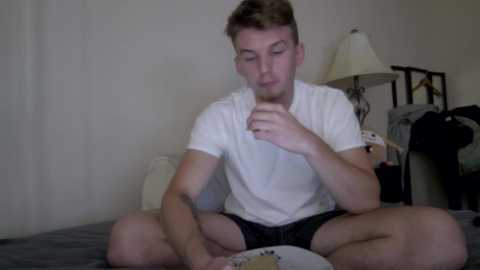 Video of a young Caucasian man with short brown hair, wearing a white t-shirt and shorts, sitting cross-legged on a bed, holding a spoon and eating a bowl of cereal.